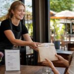 Doggy bag obligatoire en restauration : ce qu’il faut savoir A close-up shot of a smiling restaurant server handing a neatly packaged doggy bag to a satisfied diner at a cozy café table, with a sunny o