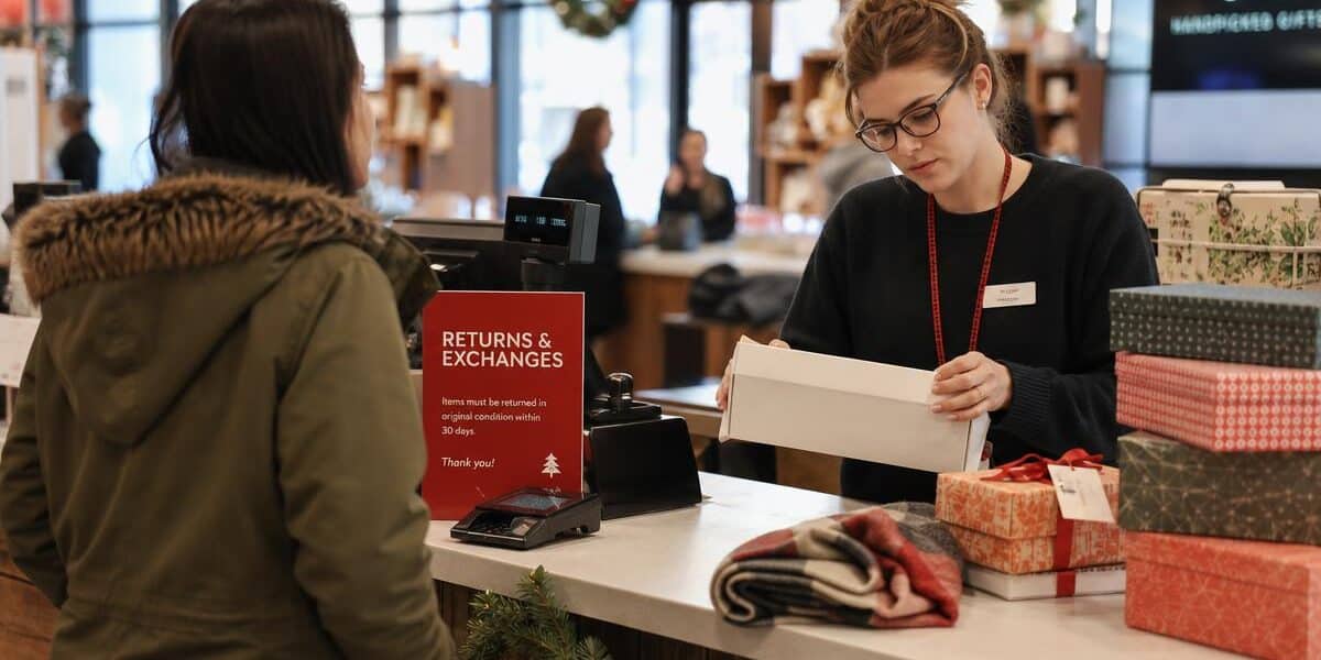 A retail employee stands at a returns counter in a bustling store, surrounded by stacks of unopened gift boxes and holiday decorations, care