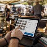 A close-up of a vendor's hands tapping on a tablet displaying a cash register software interface, surrounded by colorful second-hand items a