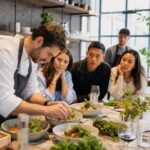 Nouveaux concepts de restauration en 2026 A chef in a modern, open kitchen is plating a vibrant, innovative dish with fresh ingredients, while a group of intrigued diners watch eager