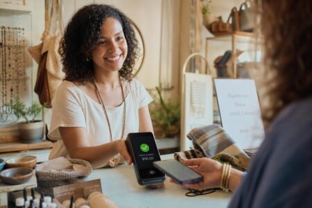 A close-up shot of a shopkeeper smiling as they assist a customer using a contactless payment terminal, surrounded by vibrant merchandise in